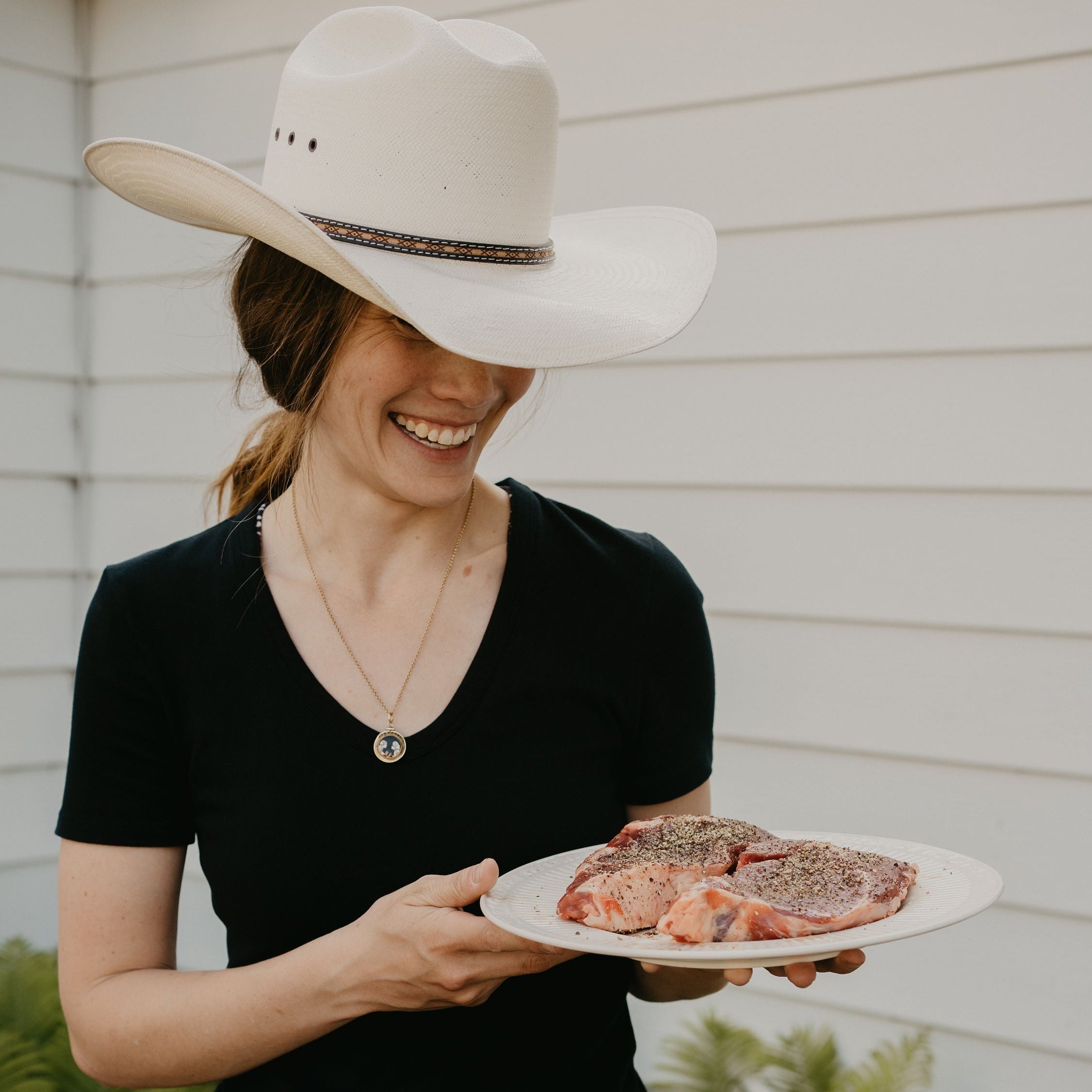 A woman wearing a white cowboy hat, black t-shirt, and blue jeans holds a white plate with raw beef steaks outdoors in front of a white house siding.