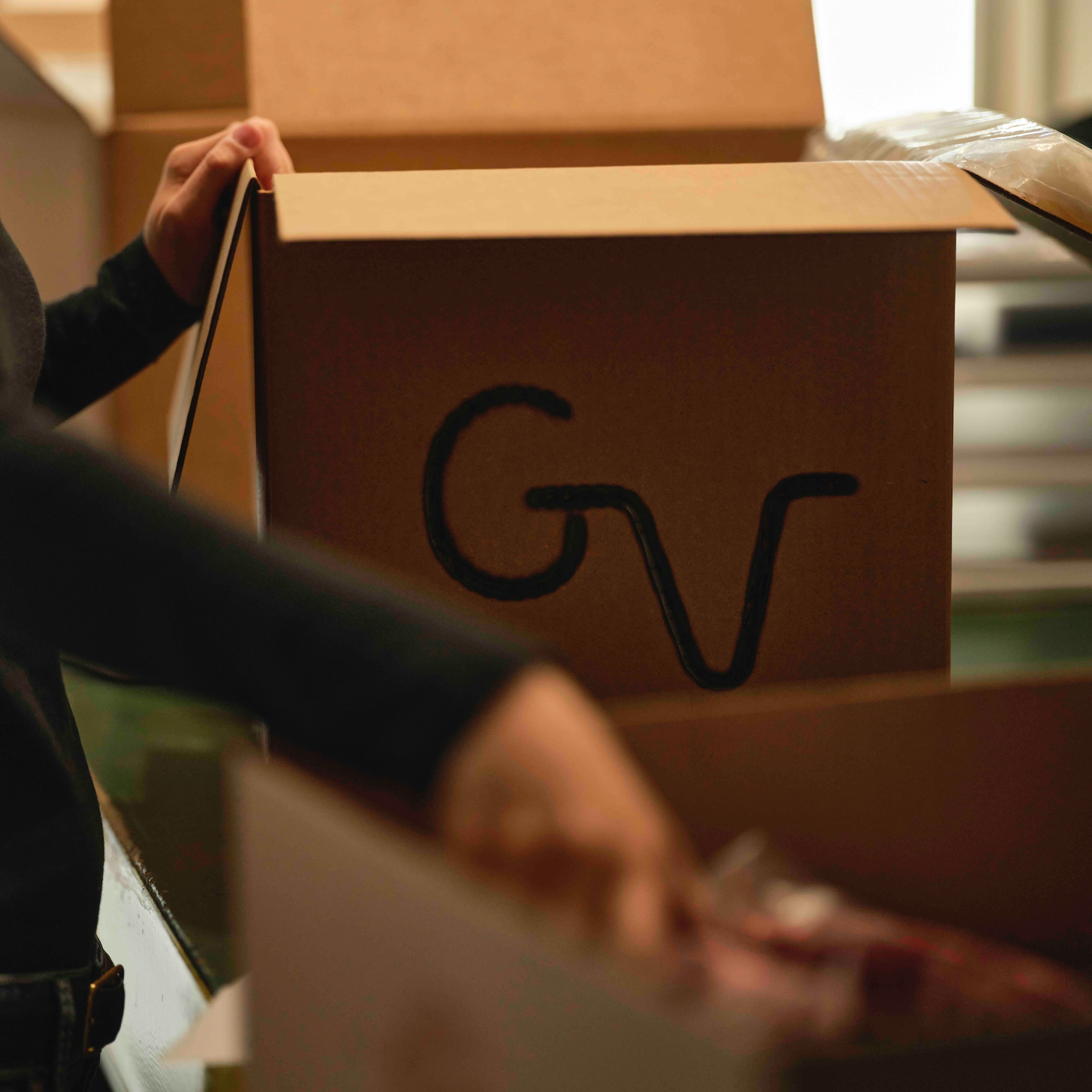 A person in a black long-sleeve top is packing or organizing items inside a large brown cardboard box with a stylized black 'GV' logo printed on the side.
